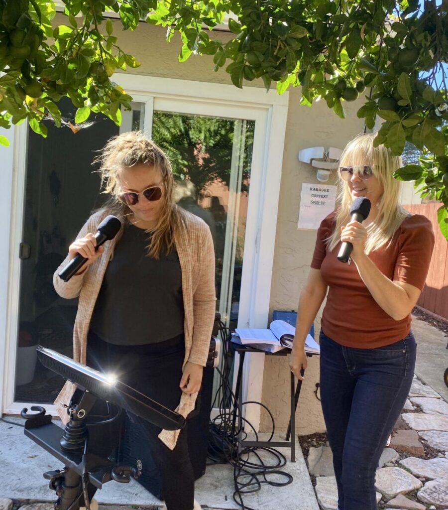 Two women standing outdoors under a tree, one is about to sing into a microphone while the other observes.