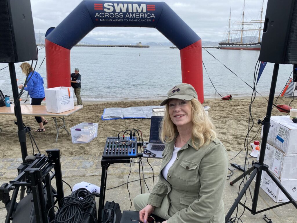A woman in a green jacket and cap sitting in front of audio equipment at a "swim across america" charity event near a body of water.