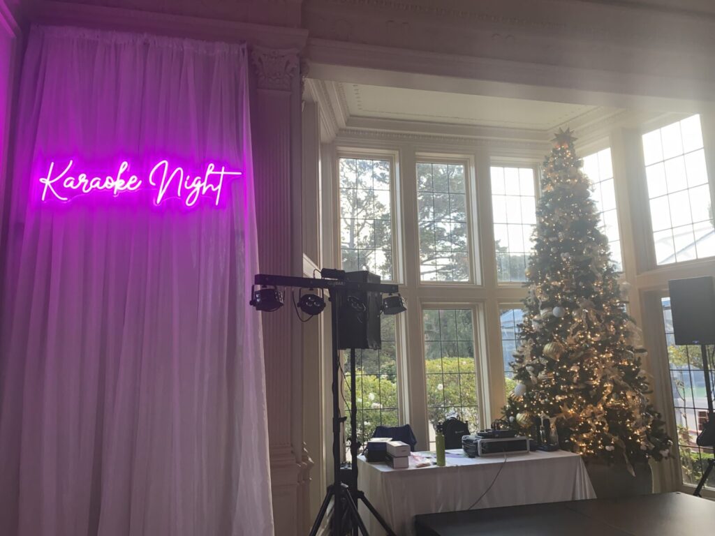 A karaoke night setup featuring a neon sign, dj equipment, and a decorated christmas tree in a room with large windows.